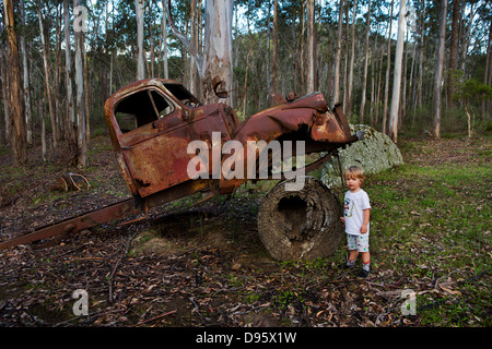 Small boy stands by the rusting cabin of an old truck propped on a tree trunk Stock Photo