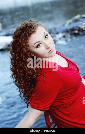Beautiful young woman with curly red hair and red dress smiles at camera with vivid blue river in background Stock Photo