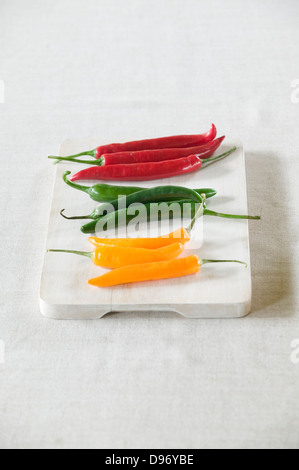 Three red, three green and three yellow chilli peppers on a white wooden chopping board. Stock Photo