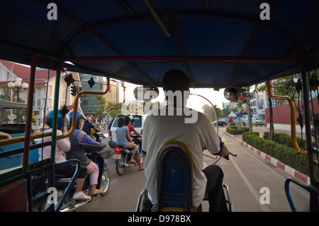 Horizontal view of a local Lao man driving a rickshaw or tuk-tuk down a busy main road in Vientiane. Stock Photo