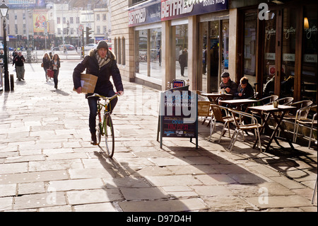 A man cycles on the pavement past a cafe in Bristol, UK, while carrying a box. Stock Photo