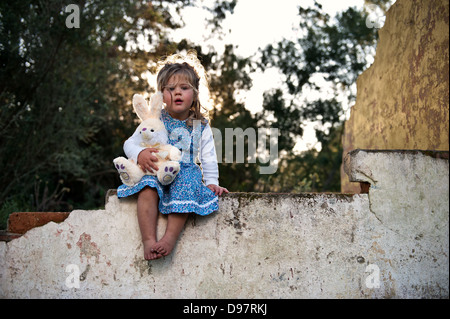 Young toddler with flowery dress and bunny teddy bear under her arm sitting on old wall of dilapidated building at sunset, cute! Stock Photo