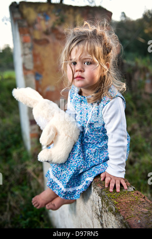 Young toddler with long blonde hair and flowery dress sitting on a wall of old dilapidated building holding a teddy bear. Stock Photo
