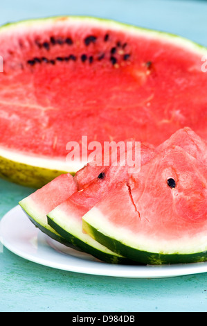 Freshly slices of pink watermelon on a plate in front of a halved melon on a blue wood surface. Stock Photo