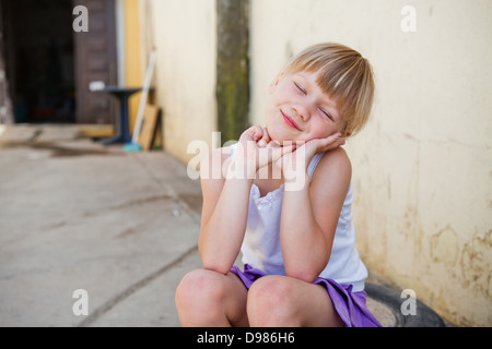 Portrait of cute smiling young girl with eyes closed sitting in front of concrete wall outside Stock Photo