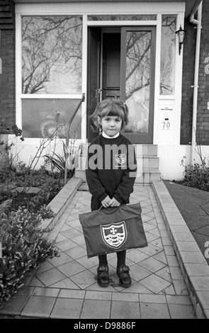 A 5 year-old girl stands outside her south London home on the first day of proper school, a momentous day and a rite of passage. Standing on the path by the front door of an Edwardian period south London home, the girl holds a brand new book bag with the initials of her local school of St Saviour's, repeated on her school jumper. She looks calm but is inwardly nervous of the day about to unfold - a rite of passage for every schoolchild, climbing the ladder of life. Stock Photo