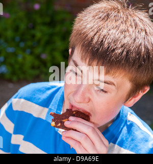 A 13 year old teenage boy eating a chocolate cookie outside in the Uk Stock Photo