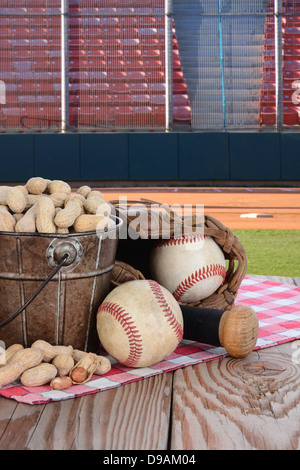 A bucket of peanuts and baseball equipment on a wood picnic table with a field and stadium in the background. Stock Photo