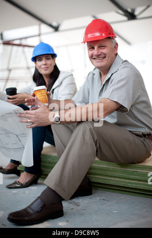 Smiling male and female construction workers sitting down holding coffee and a blueprint Stock Photo