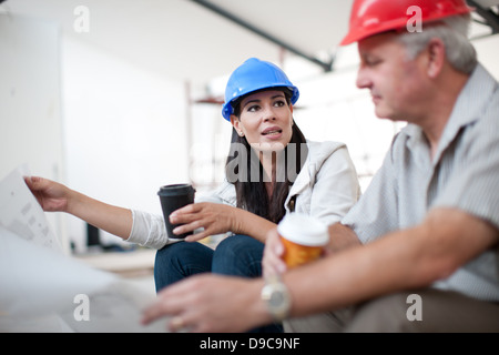 Male and female construction workers sitting down talking and holding coffee and a blueprint Stock Photo
