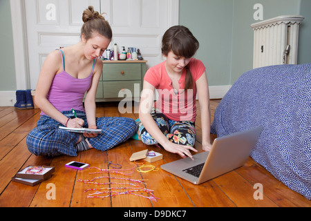 Girls sitting on bedroom floor with laptop, making friendship bracelets Stock Photo