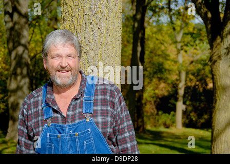 portrait of grey haired bearded farmer, wearing bib overalls & plaid shirt in meadow Stock Photo