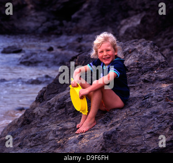 A four year old girl, on her summer holiday, posing for the camera whilst sitting on a large rock near the sea, in Cornwall, England, UK. Stock Photo