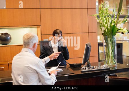 Receptionist talking on a landline phone with a businessman pointing at the hotel reception counter Stock Photo