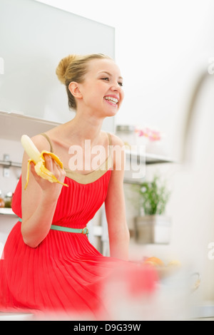Smiling young woman eating banana in kitchen Stock Photo