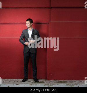 Handsome young Chinese business man leaning against a red wall. Happy Asian business man smiling and looking away. Stock Photo