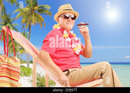Gentleman smoking a cigar and enjoying on a sun lounger on a tropical beach next to a sea and palm trees Stock Photo