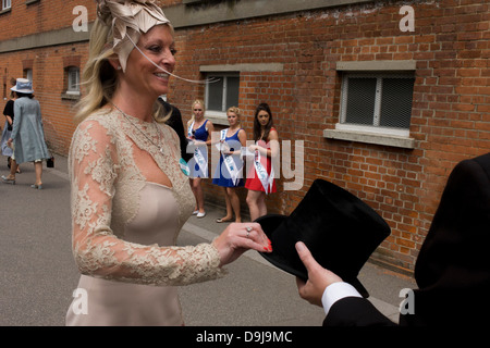 With betting promo girls looking on, a lady partner of an unseen gentleman dressed in formal menswear, passes back his top hat during the annual Royal Ascot horseracing festival in Berkshire, England. Royal Ascot is one of Europe's most famous race meetings, and dates back to 1711. Queen Elizabeth and various members of the British Royal Family attend. Held every June, it's one of the main dates on the English sporting calendar and summer social season. Over 300,000 people make the annual visit to Berkshire during Royal Ascot week, making this Europe’s best-attended race meeting with over £3m Stock Photo