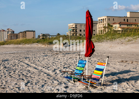 Colorful striped folding beach chairs and folded red umbrella with buildings in the background on Jacksonville Beach in Florida. Stock Photo