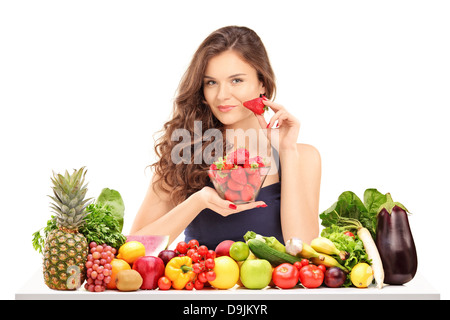Young smiling female holding a bowl of strawberries and posing behind a table full of vegetables and fruits Stock Photo