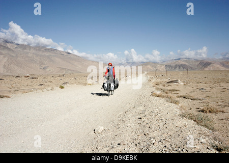 Foreign female tourist riding on a fully loaded touring bicycle on the dirt road in Pamir mountain, Tajikistan, Central Asia Stock Photo