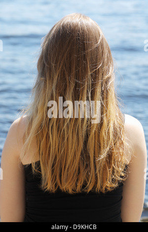 Girl with blond long hair sitting near the water back to the viewer Stock Photo