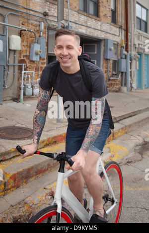 Young tattooed man cycling on street, smiling Stock Photo