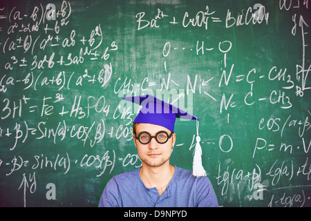 Portrait of handsome student in graduation hat and eyeglasses looking at camera on background of chalkboard Stock Photo