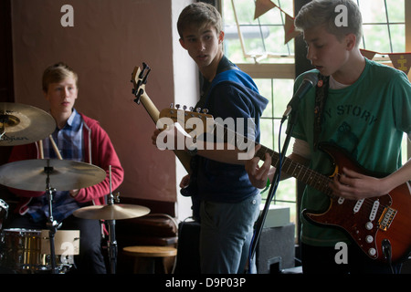 A teenage band of drums, bass and lead guitar perform in front of parents in an upstairs pub room in south London. 15 year-old lads play their own songs and covers by other musical artists. Looking over his shoulder, the bass player checks rhythm with the lead singer  and the drummer looks across his cymbals to ensure the tempo is right. The gig is a regular showcase organiused by their guitar teacher to demonstrate their musical skills as songwriters and musicians. Stock Photo