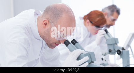 close-up of man in chemistry lab analyzing under microscope and another three researchers on the background Stock Photo