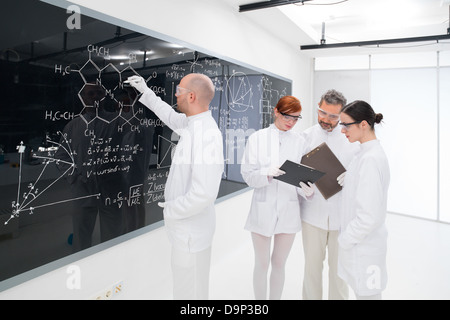 close-up of one scientist in a chemistry lab writing formulas on a blackboard and another three analyzing results on a notebook Stock Photo
