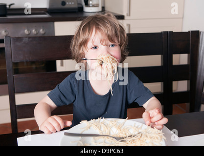 young cute boy eats spaghetti sitting on a table in the kitchen Stock Photo