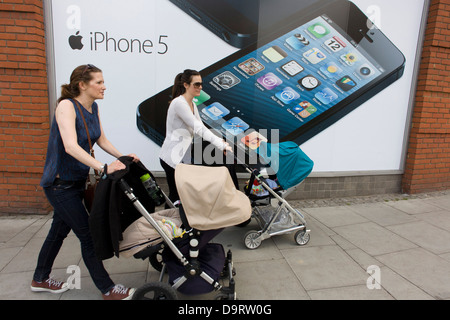 Two mothers pushing their baby's buggies, walk past a large poster for the iPhone 5 on the wall of a 'Carphone Warehouse' retailer. With the large image of its screen of this popular smartphone over their shoulders, the ladies make their way uphill in Wimbledon, London, passing this store selling Apple and other brands for consumers. Stock Photo