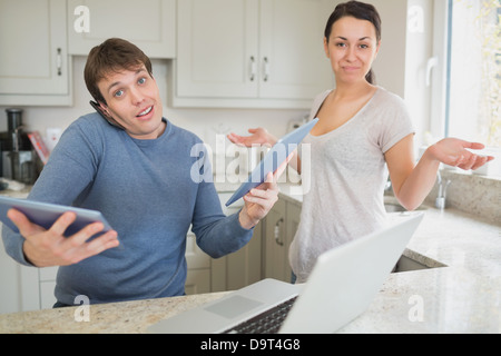 Busy man using two tablets and laptop with wife holding hands up questioning Stock Photo
