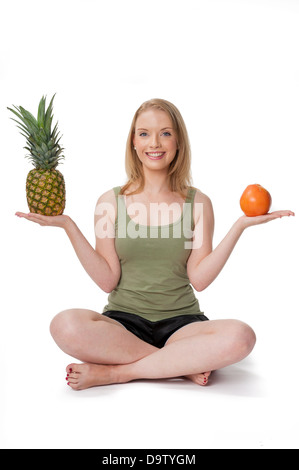 Happy young woman sitting cross-legged, holding pineapple and orange - healthy eating concept, isolated on white Stock Photo
