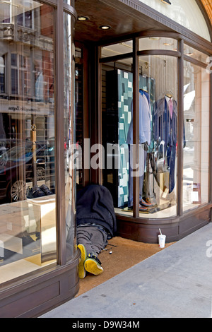 A homeless person is slumped in a doorway of an upmarket clothes store. Portrait view. Stock Photo