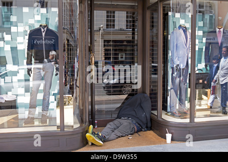 A homeless person is slumped in a doorway of an upmarket clothes store. Landscape view. Stock Photo