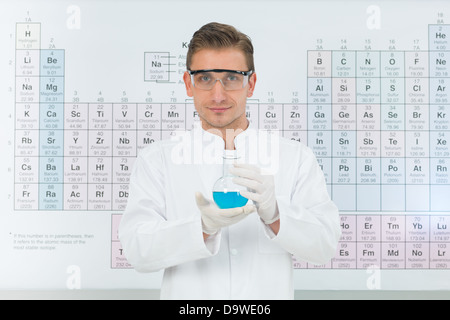 close-up of handsome male scientist smiling and holding a flask with blue substance, with periodic table in background, in a laboratory Stock Photo