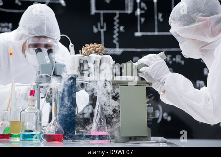 Laboratory scientists in sterile garb wearing masks and safety goggles working at a bench with microscopes surrounded by flasks Stock Photo