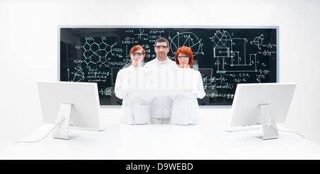 Three lab technicians dressed in white with two monitors against a blackboard with formulas and diagrams on it. Stock Photo