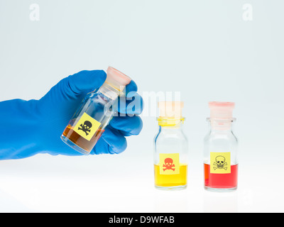 frontal view of three transparent colorless bottles filled with colored liquid and marked as poisonous, one of them held by a hand in a blue rubber glove, against a light blue background Stock Photo