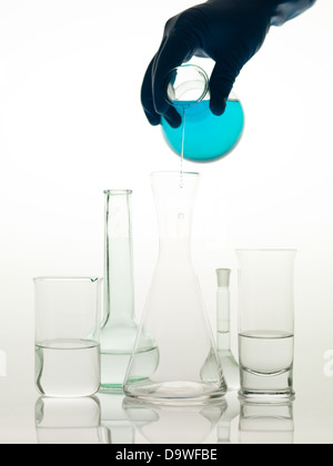 frontal view of different types of glass containers used in the laboratory, filled with transparent colorless liquid and one empty, in which someone is pouring a blue liquid Stock Photo