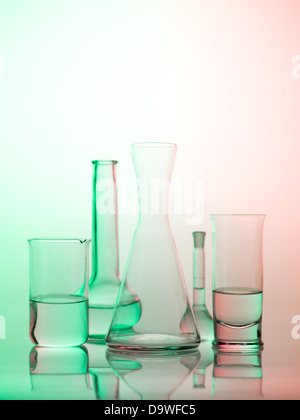frontal view of different types of glass containers used in the laboratory, filled with transparent colorless liquid and one empty, with a gradient green, red and white background Stock Photo