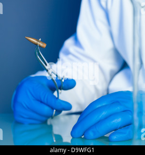 person in a white lab coat and blue rubber gloves, holding a bullet on a reflective surface Stock Photo
