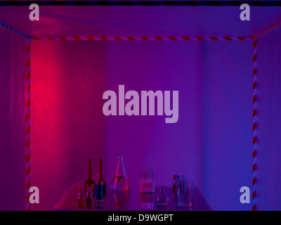 different glass containers filled with differently colored liquids on a glass table in a containment tent, lit by a gradient red, purple and blue light Stock Photo