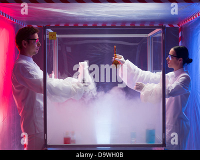 two scientists, a man and a woman, mixing chemicals in a sterile chamber labeled as bio hazardous filled with white steam, in a containment tent Stock Photo