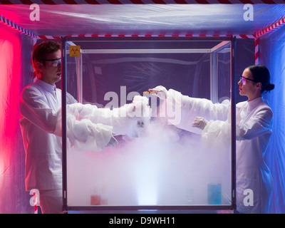 two scientists, a man and a woman, mixing chemicals in a sterile chamber labeled as bio hazardous filled with white steam, in a containment tent Stock Photo