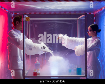 two scientists, a man and a woman, pouring substances into a glass container filled with steam and fumes, in a containment tent Stock Photo