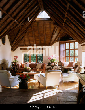 High, vaulted beamed roof in large barn conversion sitting room with white armchairs and sofa and flagstone and wooden floor Stock Photo