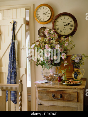 Three circular clocks on wall above rustic cupboard with vase of summer flowers in cottage dining room with apron on door Stock Photo